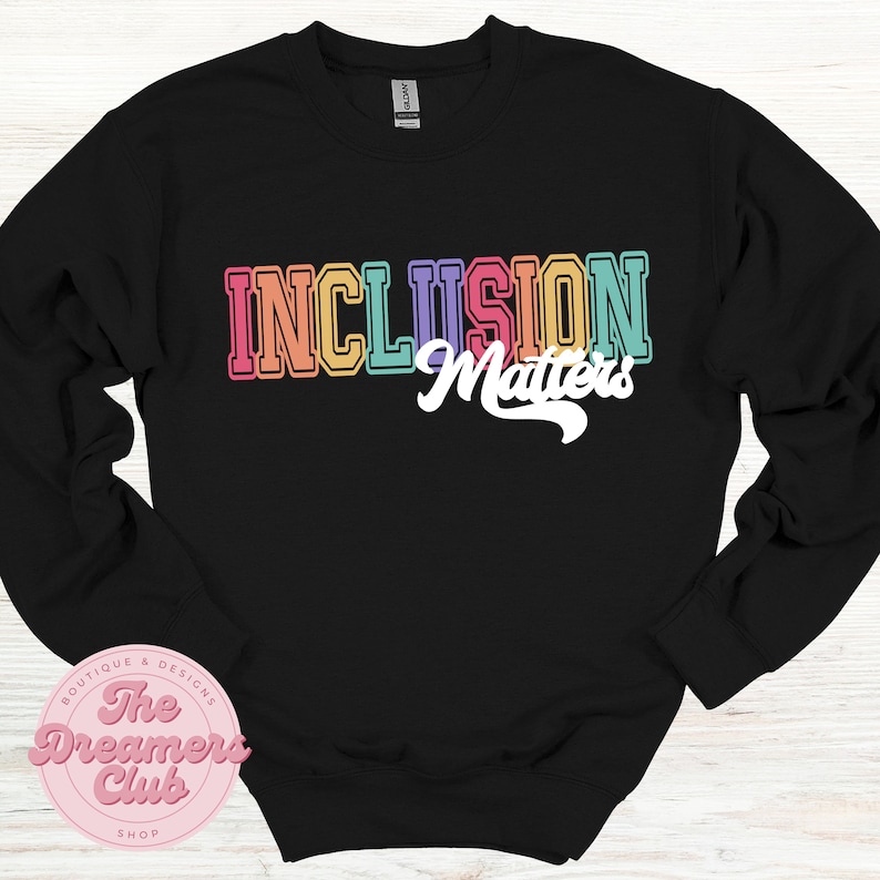 May include: Black sweatshirt with the text "INCLUSION Matters" in colorful, retro-style lettering. The words are in a rainbow of colors, including pink, orange, yellow, and blue. The sweatshirt also features a logo for "The Dreamers Club".