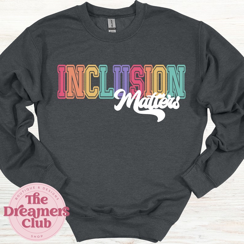 May include: Dark gray sweatshirt with the words "INCLUSION Matters" in colorful, retro-style lettering. The text is in orange, pink, purple, yellow, and teal. The sweatshirt also has a pink logo that says "The Dreamers Club Shop".