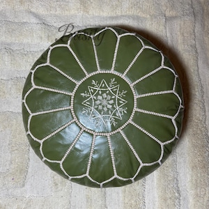 Pode incluir: Puff redondo de couro verde-oliva com costuras brancas e um desenho central de estrela bordada. Esta peça de mobiliário decorativa é adequada para uso como apoio para os pés ou assento adicional.