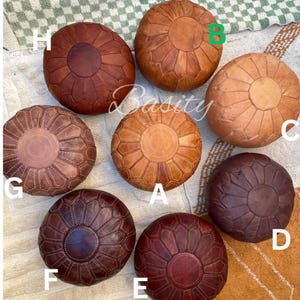 May include: Ten round leather poufs in various shades of brown, tan, and burgundy. Each pouf has a circular design with stitching around the edges. The poufs are arranged in a circle on a white and brown rug.