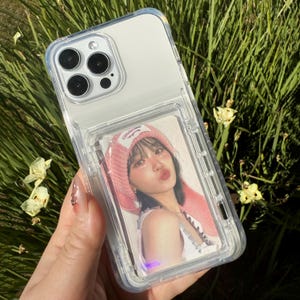 以下が含まれることがあります： ピンクの帽子をかぶった人の写真が入った透明な携帯電話ケース。携帯電話ケースは透明な背面と透明なフレームを備えています。携帯電話ケースは手で持たれています。