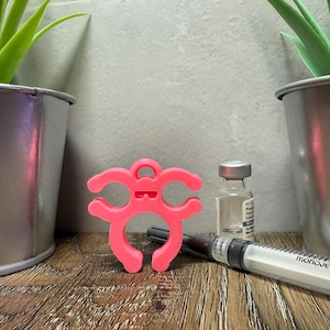 May include: A pink 3D printed  device with two loops and a small handle on top. The device is designed to hold a vial or syringe. The device is sitting on a wooden surface next to a clear glass vial and a syringe labeled "monoject".