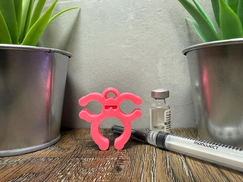 May include: A pink 3D printed  device with two loops and a small handle on top. The device is designed to hold a vial or syringe. The device is sitting on a wooden surface next to a clear glass vial and a syringe labeled "monoject".
