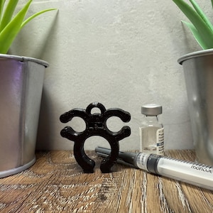 May include: A black plastic bottle stopper with a unique design. The stopper is shaped like a circle with three arms extending out. The stopper is sitting on a wooden surface next to a clear glass bottle and a syringe.
