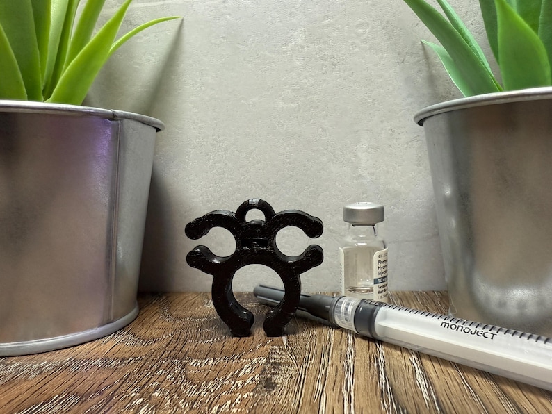 May include: A black plastic bottle stopper with a unique design. The stopper is shaped like a circle with three arms extending out. The stopper is sitting on a wooden surface next to a clear glass bottle and a syringe.