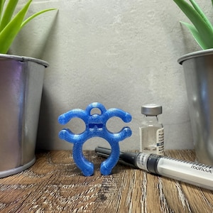 May include: A blue 3D printed syringe holder with a smiling face. The holder is designed to help secure a syringe to a surface. The holder is sitting on a wooden surface next to a syringe and a vial.