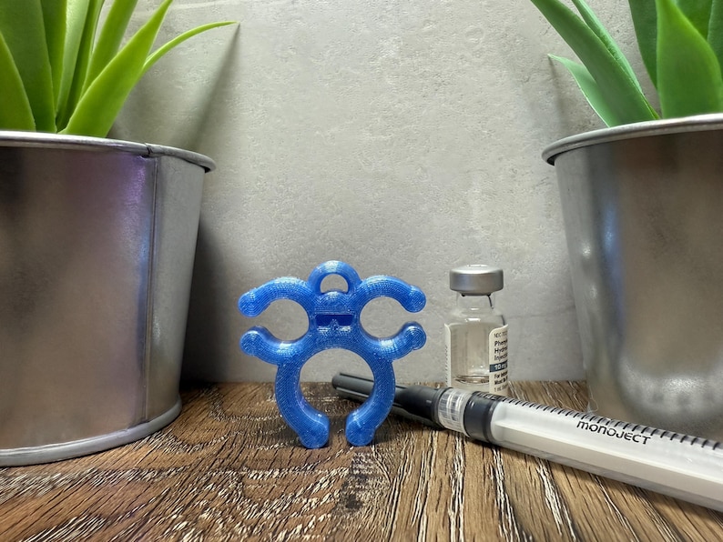 May include: A blue 3D printed syringe holder with a smiling face. The holder is designed to help secure a syringe to a surface. The holder is sitting on a wooden surface next to a syringe and a vial.