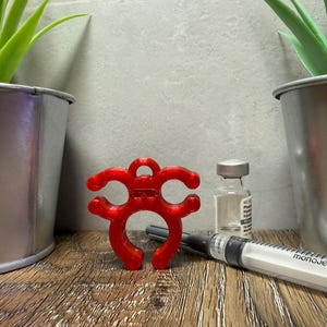 May include: A red 3D printed bottle stopper with a unique design. The stopper is shaped like a stylized creature with multiple loops. The stopper is sitting on a wooden surface next to a clear glass vial and a syringe labeled "Monoject".
