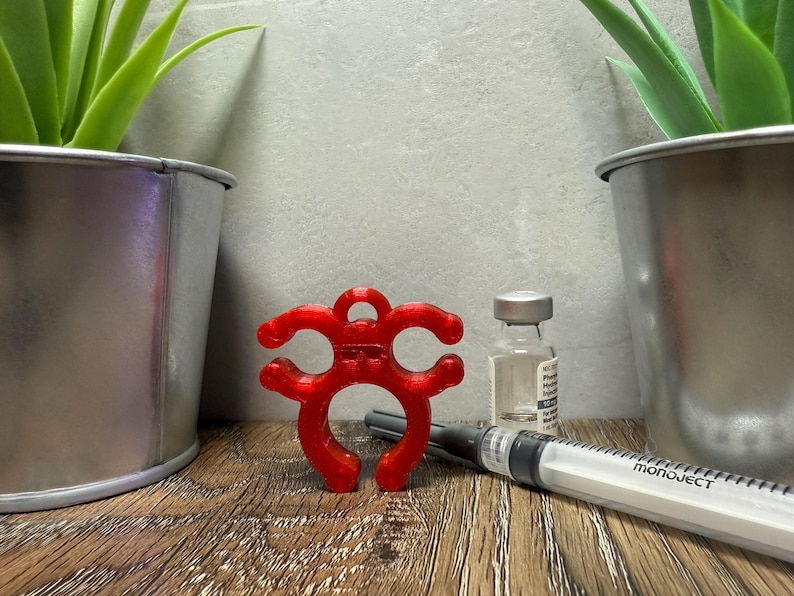 May include: A red 3D printed bottle stopper with a unique design. The stopper is shaped like a stylized creature with multiple loops. The stopper is sitting on a wooden surface next to a clear glass vial and a syringe labeled "Monoject".