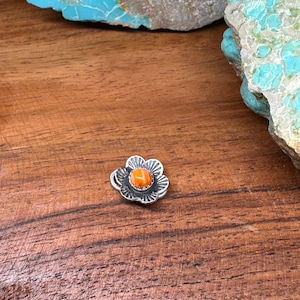 May include: A silver-toned flower-shaped pendant with an orange center stone. The pendant is set against a wooden surface, with turquoise-colored rocks in the background. The flower has detailed petals.