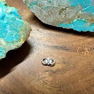 May include: A silver infinity symbol charm on a wooden surface with turquoise stones.