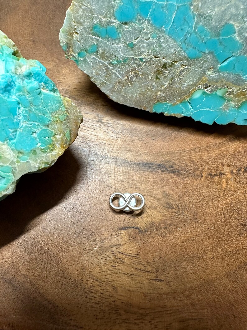 May include: A silver infinity symbol charm on a wooden surface with turquoise stones.