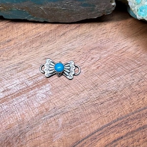 May include: A small, silver-colored bow-shaped charm with a turquoise-colored stone in the center. The charm has decorative etched details and two loops on either side. The charm is displayed on a wooden surface.
