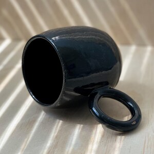 May include: A black ceramic mug with a rounded body and a large, circular handle. The mug is sitting on a light wood surface.