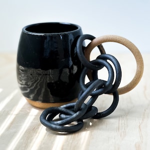 Puede incluir: Una taza de cerámica negra con un asa única hecha de una cadena negra y un anillo beige. La taza tiene un acabado brillante y una base texturizada.