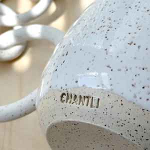 May include: White ceramic mug with speckled glaze and a brown handle. The mug has the word "CHANTLI" stamped on the bottom.