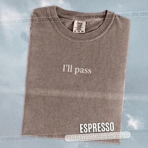 May include: Espresso-colored t-shirt with the text "I'll pass" in white. The shirt is a Comfort Colors brand, size XL-TG-EG. The t-shirt is folded and displayed on a light blue background.