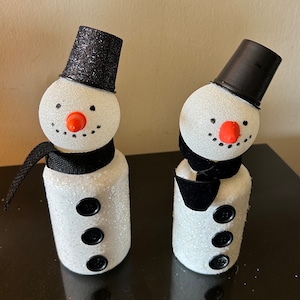 May include: Two white snowman ornaments with black hats and scarves. The snowmen have black buttons and red noses.