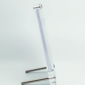 May include: A clear acrylic photo frame with two silver-colored metal support rods. The frame is designed to hold a photograph and has a minimalist, modern aesthetic. The frame is standing upright on a white surface.