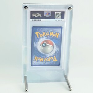 May include: A clear acrylic display stand holding a graded Pok&eacute;mon trading card. The card is encased in a protective holder with the PSA logo and a QR code. The stand has two silver metal support legs.