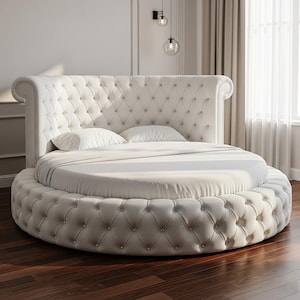 May include: A luxurious, round bed with a tufted headboard and base. The bed is upholstered in a cream-colored fabric and features a button-tufted design. Two white pillows rest on the bed, which is covered with a white sheet and blanket. The bed is set on a dark wood floor.