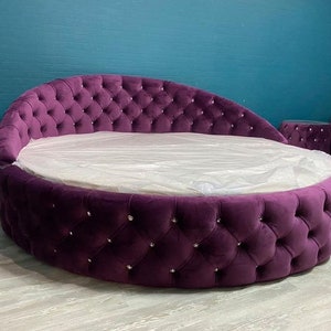 May include: A large, round, purple velvet bed with a tufted headboard and diamond-shaped buttons. The bed is covered in a white sheet and has a small, round, purple velvet ottoman next to it.
