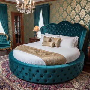 May include: A luxurious, round teal bed with a tufted headboard and base. The bed is adorned with white bedding, gold pillows, and a gold runner. The room features teal curtains, a patterned rug, and a chandelier, creating an elegant bedroom setting.