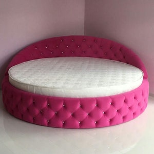 May include: A round pink bed with a white mattress and diamond-shaped tufting. The bed has a curved headboard and is upholstered in a soft, plush fabric.