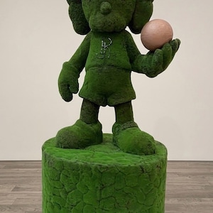 May include: A green sculpture of a cartoon character with X-shaped eyes, holding a pink sphere. The figure is standing on a green, textured pedestal. The sculpture is covered in a moss-like material.
