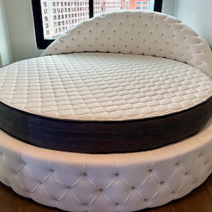 May include: A white round bed with a tufted headboard and a gray mattress. The bed is in a modern bedroom with a window in the background.