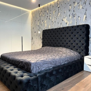 May include: A black tufted upholstered bed with a gray blanket on top. The headboard is tall and has a tufted design. The bed is in a bedroom with a white wall and a gray textured wall behind the bed.