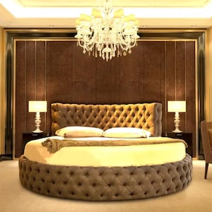 May include: A round, tufted bed with a brown headboard and a gold frame. The bed is in a bedroom with a chandelier and two lamps.