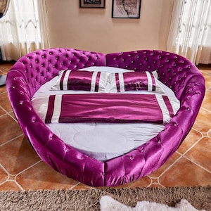May include: A large, round, purple velvet bed with a tufted headboard and diamond-shaped buttons. The bed is covered in white linens and has two white pillows with purple stripes.