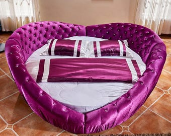 May include: A large, round, purple velvet bed with a tufted headboard and diamond-shaped buttons. The bed is covered in white linens and has two white pillows with purple stripes.