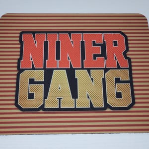 May include: Rectangular mousepad with the words "NINER GANG" in orange and yellow block letters. The text is outlined in black and set against a striped background of red and tan.