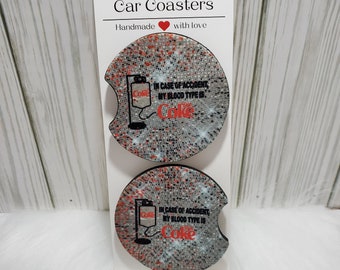 May include: Two round car coasters with a white background and red and black speckles. The coasters feature a graphic of a Coca-Cola bottle with an IV drip and the text "IN CASE OF ACCIDENT, MY BLOOD TYPE IS Coke".