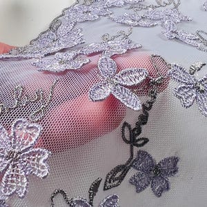 May include: Close-up of delicate lavender lace fabric with embroidered floral designs. The lace features intricate details, including silver metallic thread accents and a scalloped edge. The fabric is held up, revealing a sheer, net-like backing.