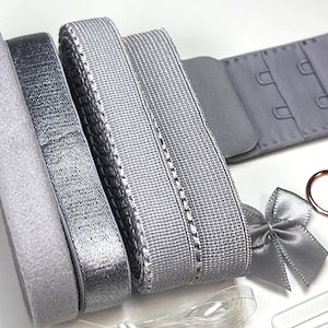 May include: A selection of grey bra-making components, including elastic, hook and eye fastenings, and a small satin bow. The elastic ribbons have different textures and sheens, with a soft, velvety band and a metallic silver ribbon.