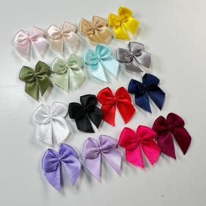 Puede incluir: Una colección de pequeños lazos de satén en varios colores, incluyendo rosa, dorado, amarillo, verde, azul, gris, negro, rojo y morado. Estos lazos decorativos están dispuestos sobre una superficie blanca, mostrando sus vibrantes colores y texturas.