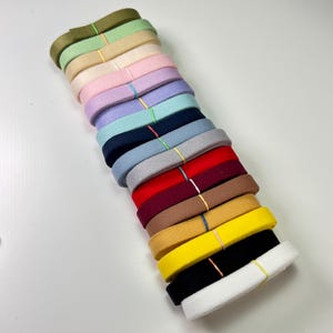 Pode incluir: Uma pilha de fitas de feltro coloridas em várias tonalidades, incluindo verde, rosa, roxo, azul, vermelho, castanho, amarelo, preto e branco. Cada fita é presa com uma fina faixa elástica. As fitas estão dispostas diagonalmente sobre um fundo branco.