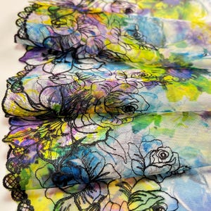 May include: Close-up of a floral fabric with a watercolor effect. The fabric features a vibrant mix of colors, including blue, yellow, purple, and green, with black floral outlines and a scalloped edge. The design is layered and detailed.