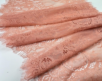 May include: Close-up of a peach-colored lace fabric with intricate floral patterns. The delicate material is folded, showcasing the detailed design and fringed edges. The lace is ideal for crafting and decorative projects.