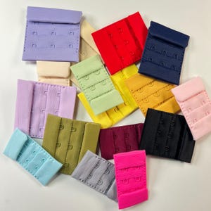 May include: A collection of colorful bra extenders in various shades, including pink, red, blue, green, and black. Each extender features multiple hook-and-eye closures for adjustable sizing. The extenders are made of fabric and metal.