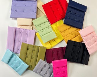 May include: A collection of colorful bra extenders in various shades, including pink, red, blue, green, and black. Each extender features multiple hook-and-eye closures for adjustable sizing. The extenders are made of fabric and metal.