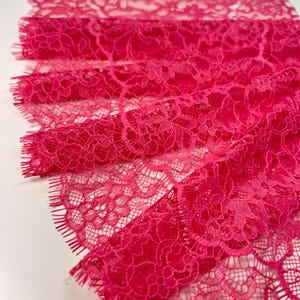 Op de afbeelding: Close-up van levendig fuchsia kanten stof. Het delicate bloemenpatroon is zichtbaar in het hele textiel. De randen van het kant zijn gefranjerd. De stof is opgerold in verschillende lagen, wat het ingewikkelde ontwerp en de textuur laat zien.