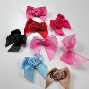 May include: A selection of decorative bows in various colours. The bows are made from satin and sheer fabrics, with shades of pink, red, black, blue, and tan. Each bow has a small rose in the centre. Ideal for craft projects and embellishments.