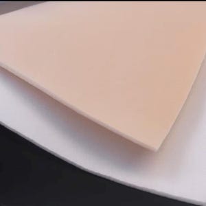 May include: A rectangular sheet of smooth, light peach-colored synthetic material layered on top of a larger, white, slightly textured fabric sheet. The peach layer has a clean, straight edge and a uniform, matte surface finish throughout.