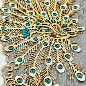 May include: An embroidered peacock feather design in gold, teal, and white. The intricate pattern features a detailed fan shape with teal accents and a delicate, lacy edge. The design is on a sheer black fabric.