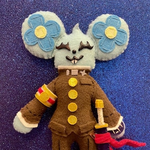May include: A handmade felt mouse doll with light blue fur, large flower-shaped ears, and a brown military-style uniform. The doll holds a purple and gold sword with a red sash. The background is a sparkly blue.