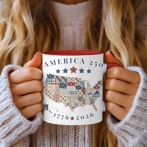 May include: White ceramic mug with a red interior and handle, featuring the text "AMERICA 250" above a patchwork map of the United States. The bottom of the mug displays "1776-2026" with stars. The mug is held by a person.